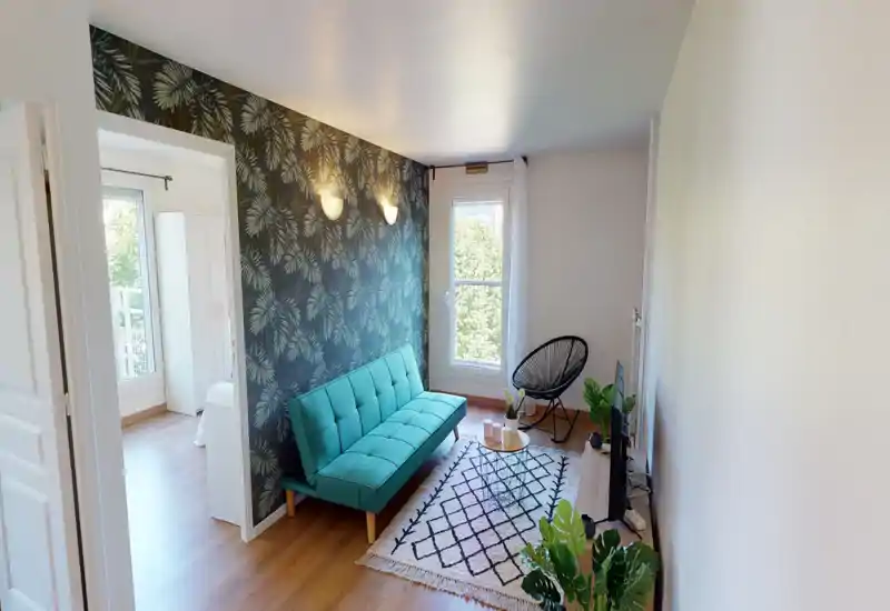 Salon lumineux et soigné avec un canapé turquoise, une décoration murale, un tapis et des plantes d'intérieur. La lumière naturelle depuis la fenêtre crée une atmosphère chaleureuse.