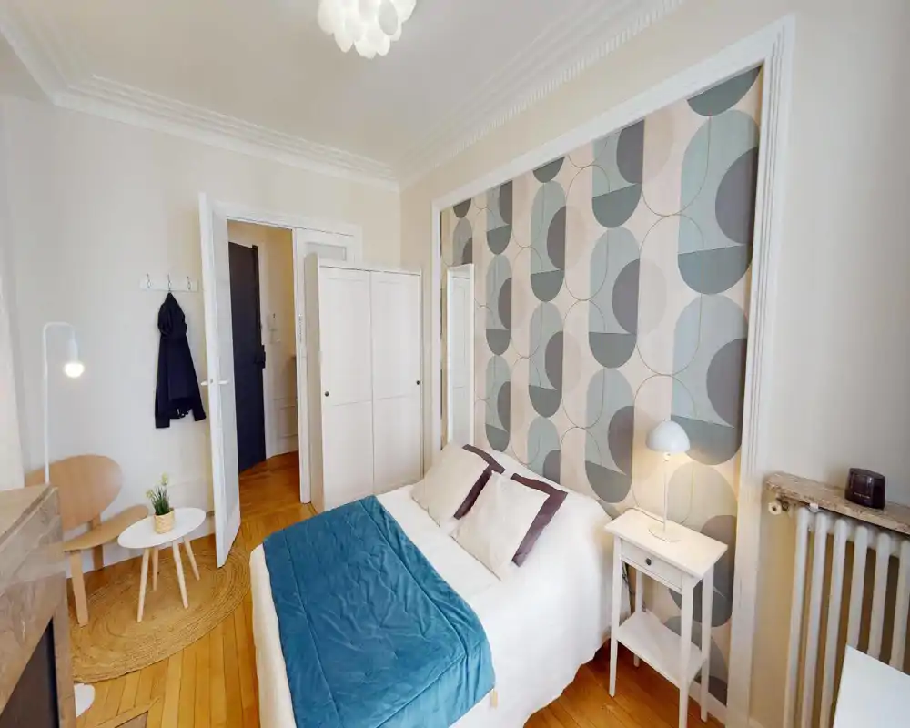 Chambre lumineuse et bien mise en scène avec lit double, papier peint géométrique et table de chevet avec lampe — idéale pour présenter un espace de nuit chaleureux.
