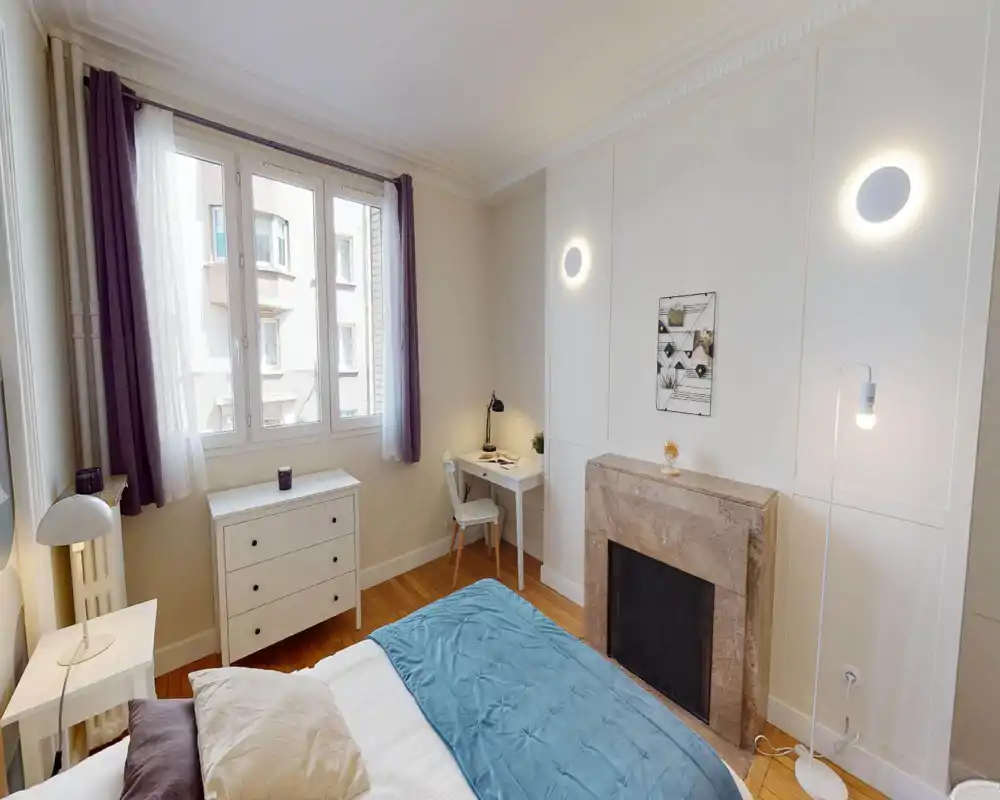 Chambre lumineuse et confortable avec grande fenêtre, lit bien fait avec jeté bleu, table de chevet et petit bureau — idéale pour se reposer et travailler occasionnellement.