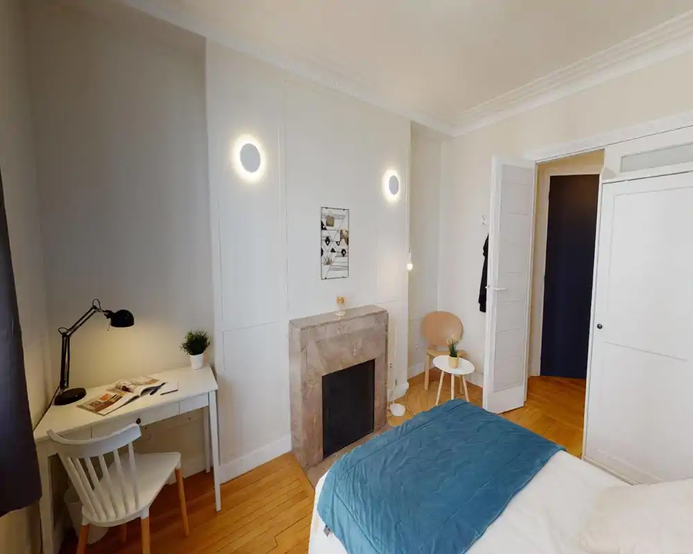 Chambre lumineuse et décorée avec lit double visible, parquet, petit bureau et une cheminée décorative qui apportent du charme.