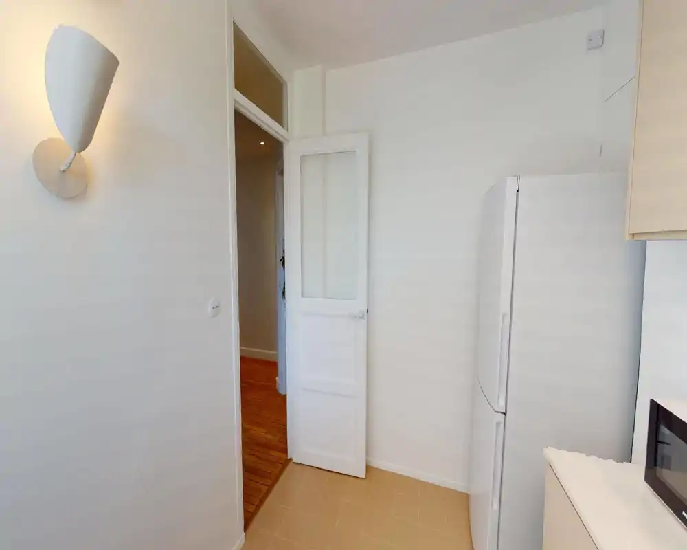 Compact, clean kitchen corner with a white refrigerator and countertop visible; neutral tiles and simple wall lighting create a functional, minimal feel.