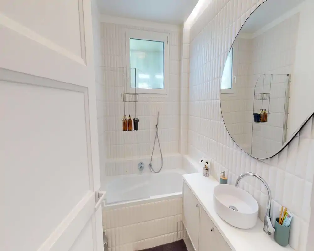 Bright, modern bathroom with a bathtub, wall-mounted shower hose, round mirror and a countertop sink—clean white tiles and compact, stylish storage.