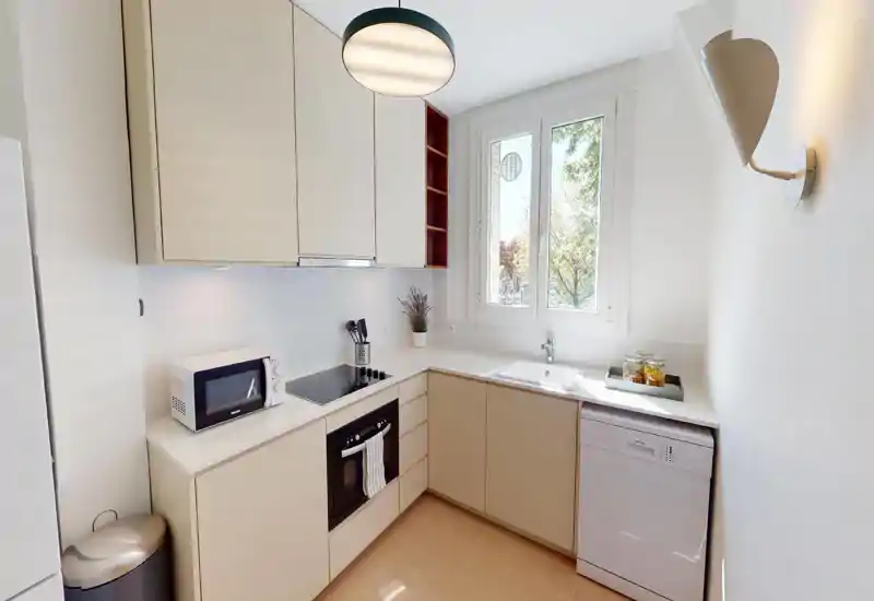 Bright, compact modern kitchen with beige cabinets, built-in oven, induction cooktop, microwave and a sink under a window letting in natural light.