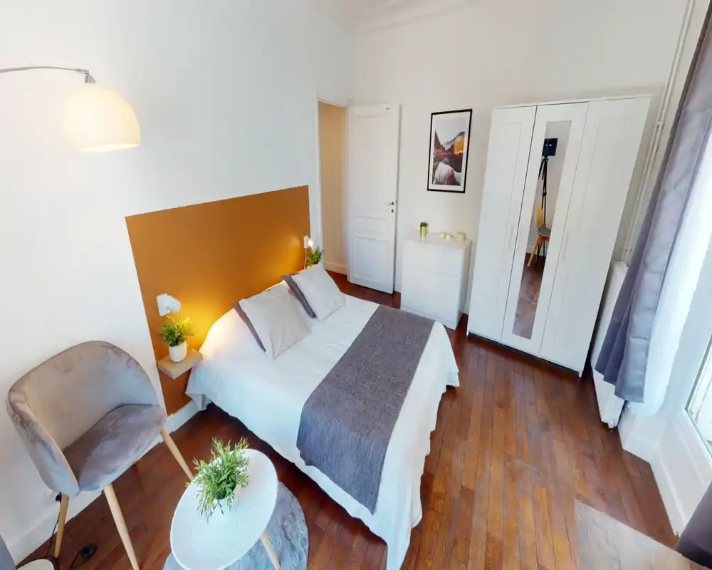 Chambre lumineuse et élégante avec lit double centré, tête de lit colorée, lampes de chevet et plantes décoratives ; parquet et armoire blanche complètent l’espace chaleureux et soigné.