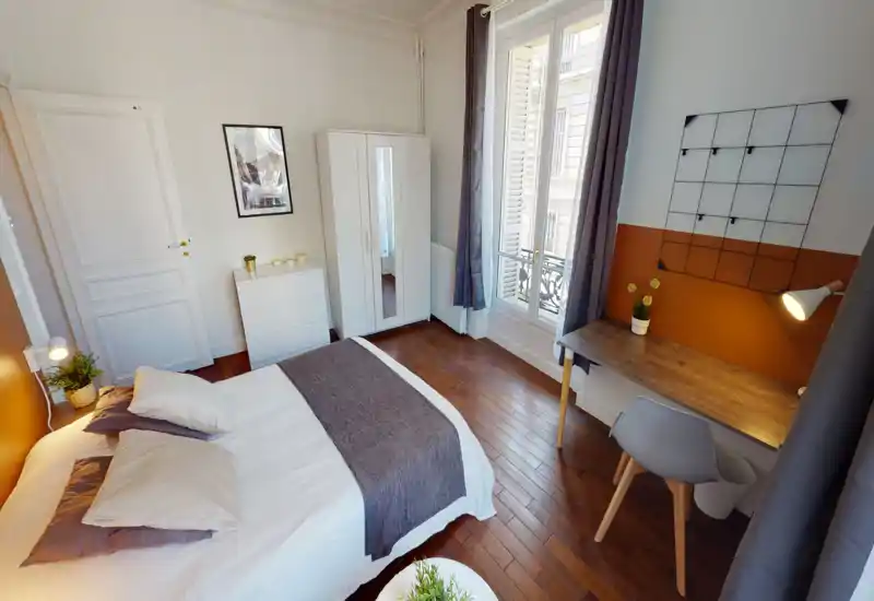 Chambre lumineuse et bien décorée avec un grand lit au premier plan, parquet, armoire et un bureau près d'une grande fenêtre laissant entrer la lumière naturelle.