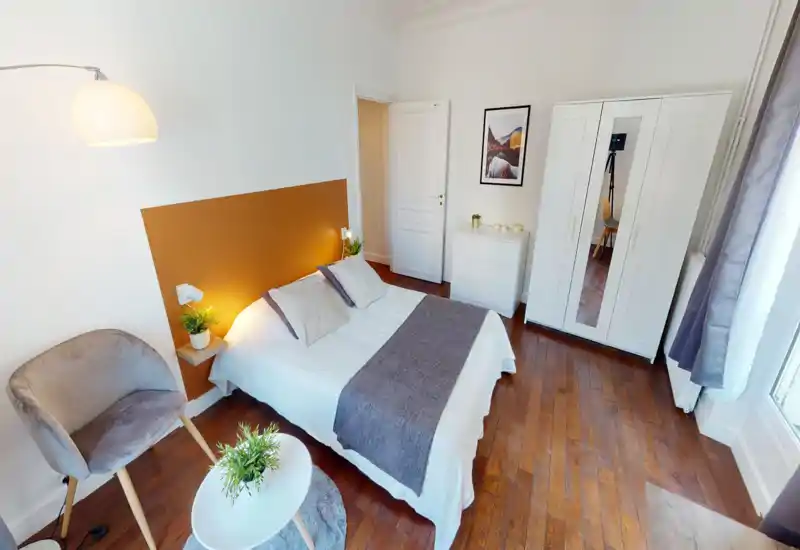 Chambre lumineuse et élégante avec lit double centré, tête de lit colorée, lampes de chevet et plantes décoratives ; parquet et armoire blanche complètent l’espace chaleureux et soigné.