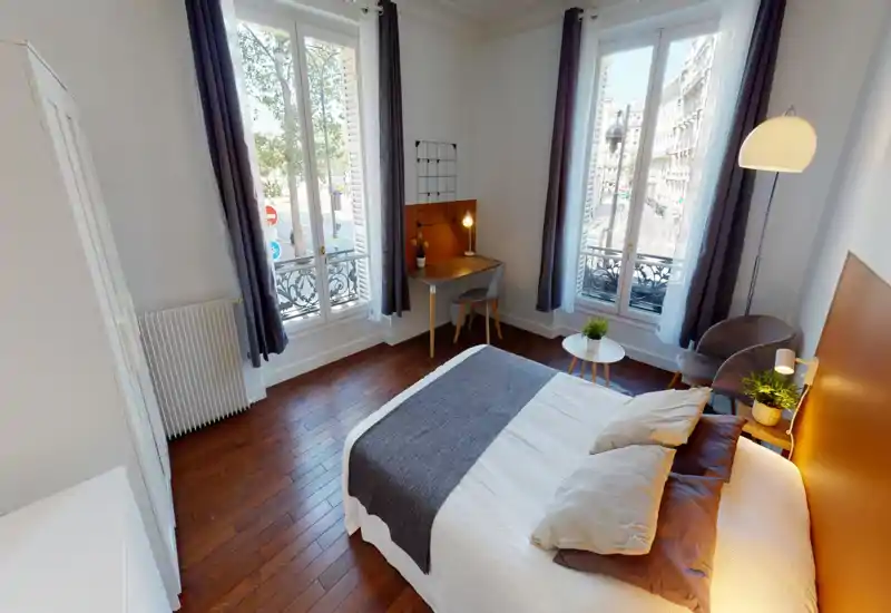 Chambre lumineuse et élégante avec un lit double confortable, parquet chaleureux et deux grandes fenêtres offrant une belle lumière naturelle et vue sur la ville. La pièce dispose d’un petit bureau, d’un coin salon et d’une décoration soignée.