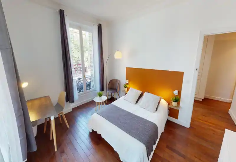Chambre double lumineuse et bien décorée avec tête de lit orange, grande fenêtre, bureau et coin sièges cosy — idéale pour un séjour confortable et lumineux.