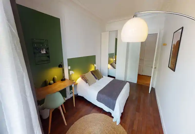 Chambre lumineuse et élégante avec un lit double habillé de linge blanc, coussins décoratifs et plaid gris. Un bureau en bois et une chaise se trouvent à côté du lit sur un mur vert ; une armoire miroir et des lampes de chevet chaudes ajoutent du confort.