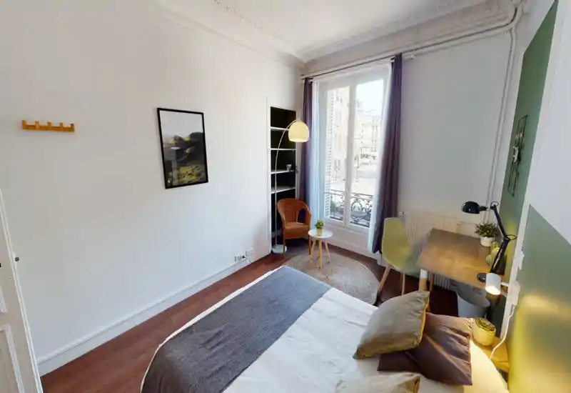 Chambre lumineuse et aménagée avec un lit au premier plan, un coin lecture cosy près d'une grande fenêtre et un petit bureau—parfaite pour un séjour en ville.