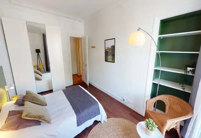 Chambre lumineuse et mise en scène, avec un lit double bien fait, des textiles doux et un coin lecture confortable avec fauteuil en osier et lampe. Placard miroir et étagères intégrées apportent rangement et profondeur, le parquet chaud rend l'espace accueillant.