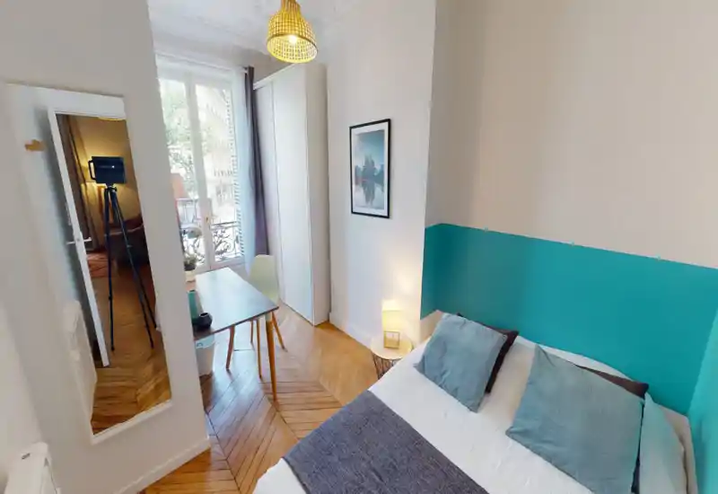 Camera da letto luminosa e accogliente con letto matrimoniale, cuscini decorativi, lampada da comodino e scrivania vicino alla finestra con vista sulla strada. Arredo moderno e pulito con parete accentuata turchese e pavimento in legno a spina di pesce.
