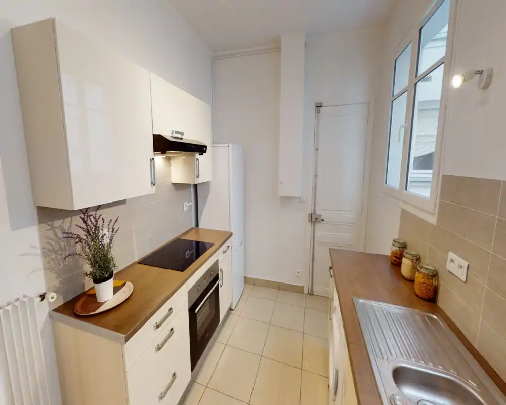 Cuisine compacte et lumineuse avec plans de travail imitation bois, four encastré et plaque vitrocéramique, réfrigérateur indépendant et évier en inox ; finitions modernes et aménagement fonctionnel.