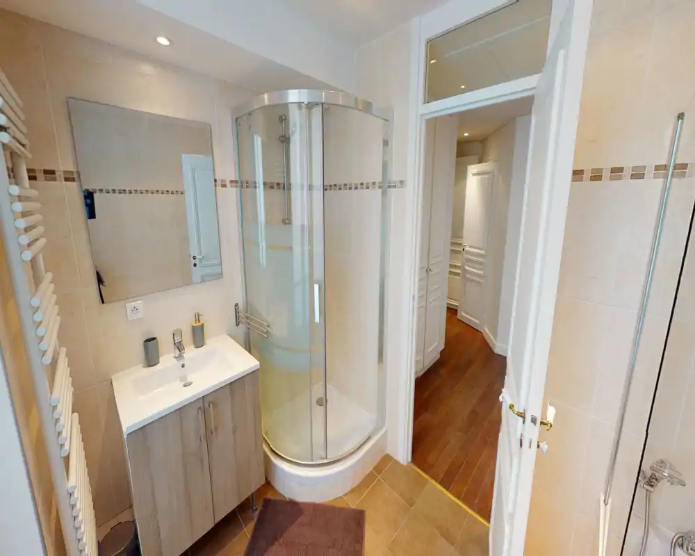 Salle de bains moderne et lumineuse avec cabine de douche d'angle en verre, meuble vasque effet bois, grand miroir mural et sèche-serviettes.