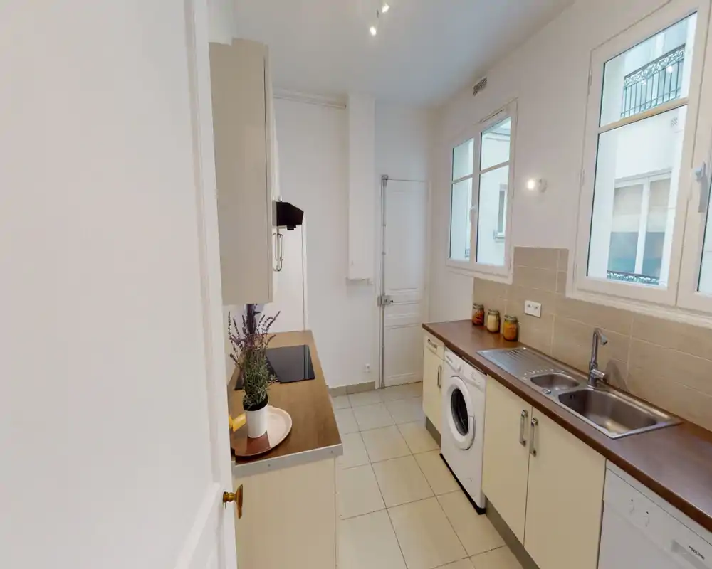 Cuisine étroite et lumineuse avec plans de travail en bois, évier intégré, machine à laver et plaque à induction ; finitions modernes et deux fenêtres apportant la lumière.