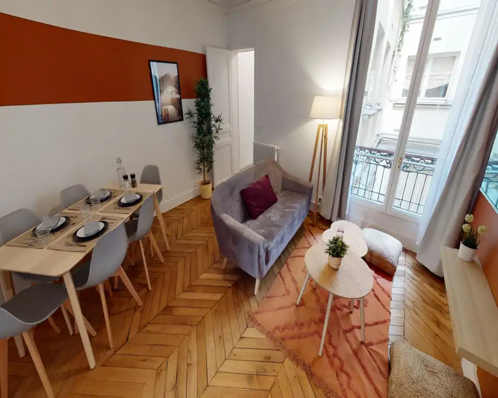 Espace de vie lumineux et aménagé avec un canapé gris confortable, tapis d'accent et table à manger près d'une grande fenêtre laissant entrer la lumière naturelle.