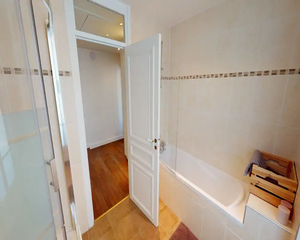 Salle de bain carrelée lumineuse avec baignoire intégrée, pare-douche en verre et caisse en bois pour serviettes — propre et bien éclairée.