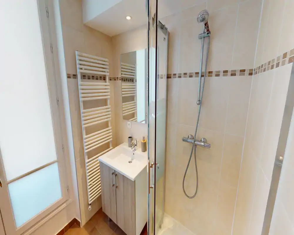 Salle de bain moderne et compacte avec cabine de douche, douchette, lavabo avec meuble et sèche-serviettes; carrelage clair et bonne luminosité.