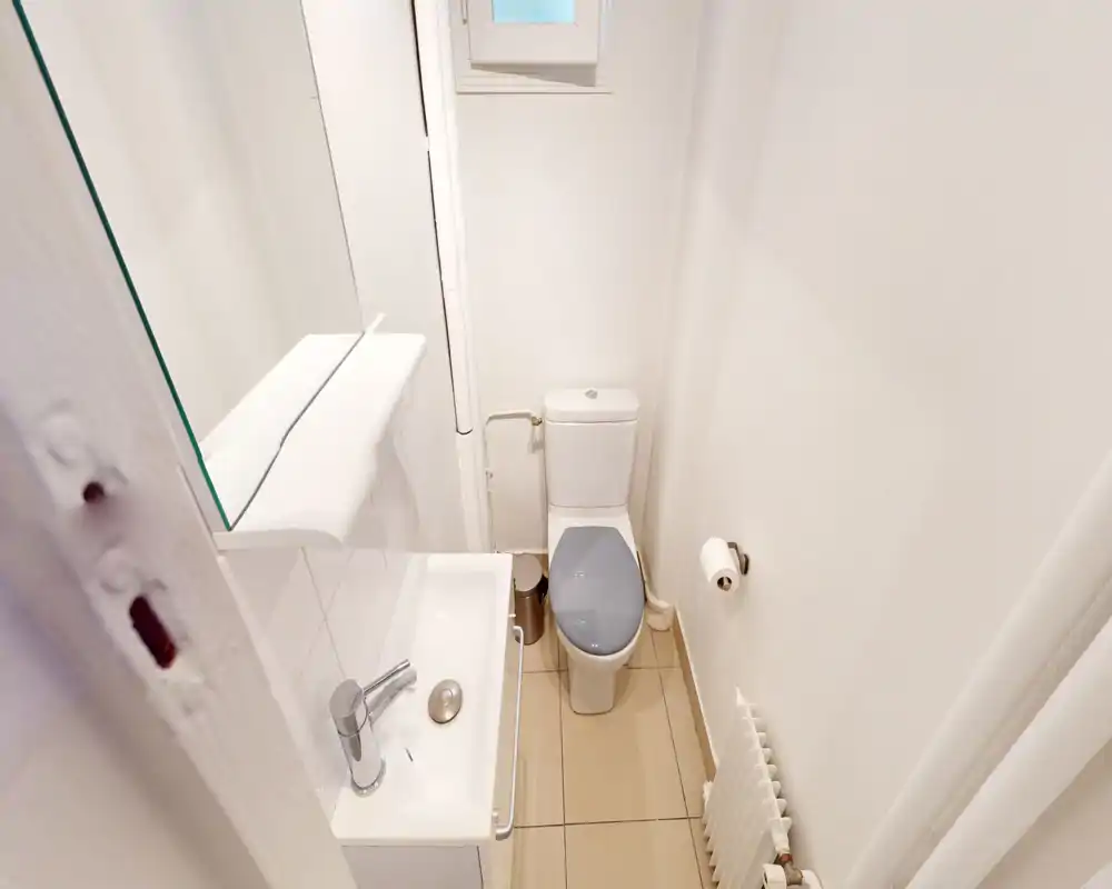 Petite salle de bain lumineuse avec lavabo, miroir et toilette. Finitions blanches et sol carrelé pour un espace propre et fonctionnel.