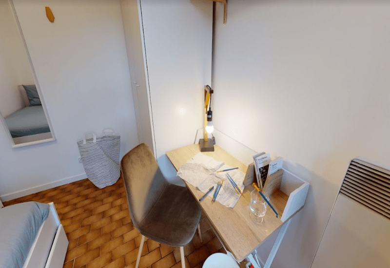 Small, tidy bedroom workspace with a single bed partially visible, a wooden desk with lamp and chair — ideal for a student or short stay.