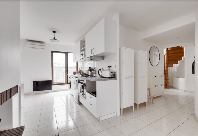 Bright modern kitchenette with white cabinetry, integrated oven and countertop appliances; clean tiled floor and a window providing natural light. The space opens toward a staircase with warm wood accents.