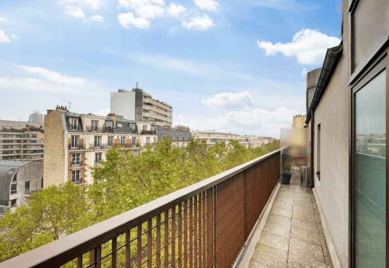 Sunny narrow balcony with a metal railing overlooking tree-lined city buildings and a clear sky — ideal for morning coffee or a small outdoor seating area.
