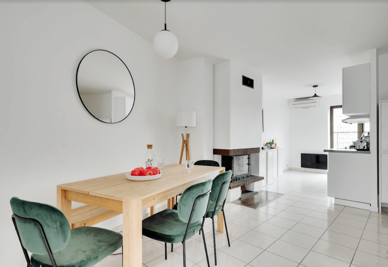 Bright, minimalist dining area with a wooden table, green velvet chairs and a decorative round mirror; open plan connects to the kitchen and living space.