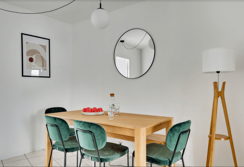 Bright, modern dining area with a wooden table, four green velvet chairs and minimalist decor including a round wall mirror and floor lamp.