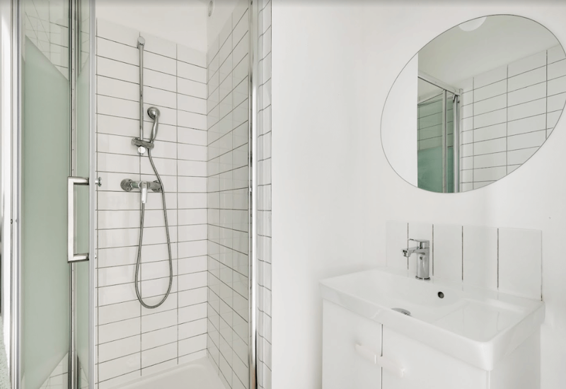 Bright modern bathroom with a tiled shower stall, chrome hand shower and a compact white vanity with round mirror — clean, minimal and well lit.