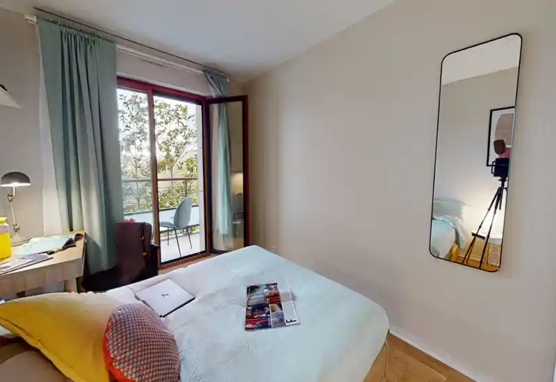 Chambre lumineuse et chaleureuse avec lit double, lampe de chevet et bureau ; porte vitrée donnant sur un petit balcon avec sièges extérieurs.