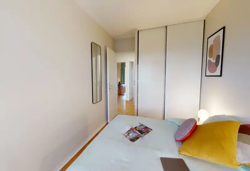 Chambre moderne et lumineuse avec lit bien fait, coussins colorés et lampe de chevet ; armoire coulissante et miroir en pied visibles, espace propre et accueillant.
