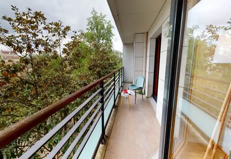 Balcone stretto e arioso con ringhiera in metallo, piccolo tavolino e sedia che si affacciano su alberi rigogliosi — perfetto per un caffè mattutino.
