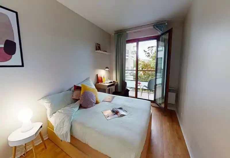 Chambre lumineuse et élégante avec grand lit double, table de chevet avec lampe, bureau près du balcon et porte-fenêtre ouverte laissant entrer la lumière naturelle.