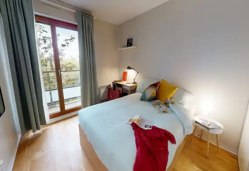 Chambre moderne et lumineuse avec lit double habillé de draps clairs, coussins colorés et lampe de chevet ; grandes portes donnant sur le balcon apportent beaucoup de lumière naturelle.
