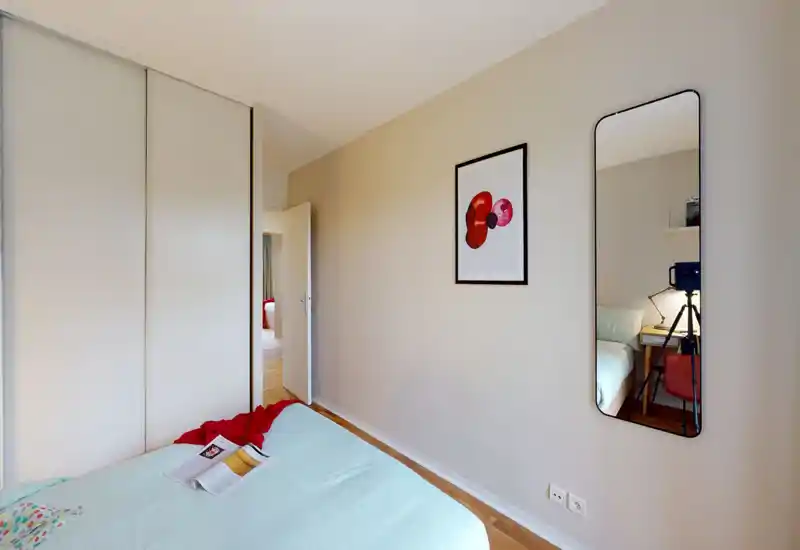 Chambre minimaliste et lumineuse avec un lit double bien fait, une literie claire et un jeté décoratif. Un miroir en pied et un cadre ajoutent de la profondeur au mur neutre.