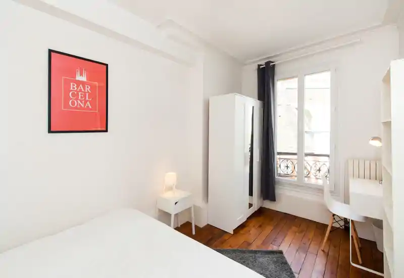 Bright, minimal bedroom with a comfortable bed, bedside table with lamp, wardrobe and a small desk by a large window letting in plenty of light.