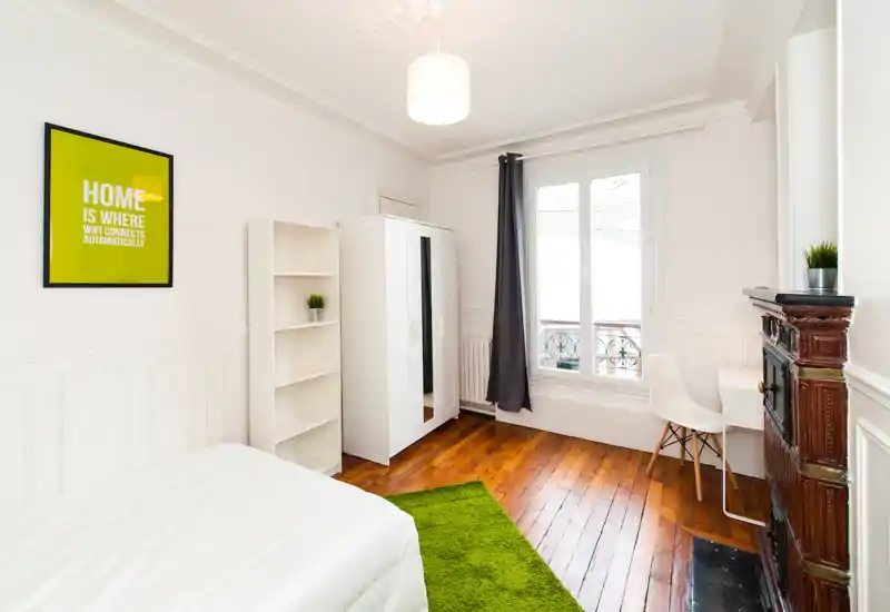 Bright, minimal bedroom with a white bed, green rug and wooden floors. The room features a wardrobe, shelving and a window with blackout curtain, creating a clean, comfortable space.