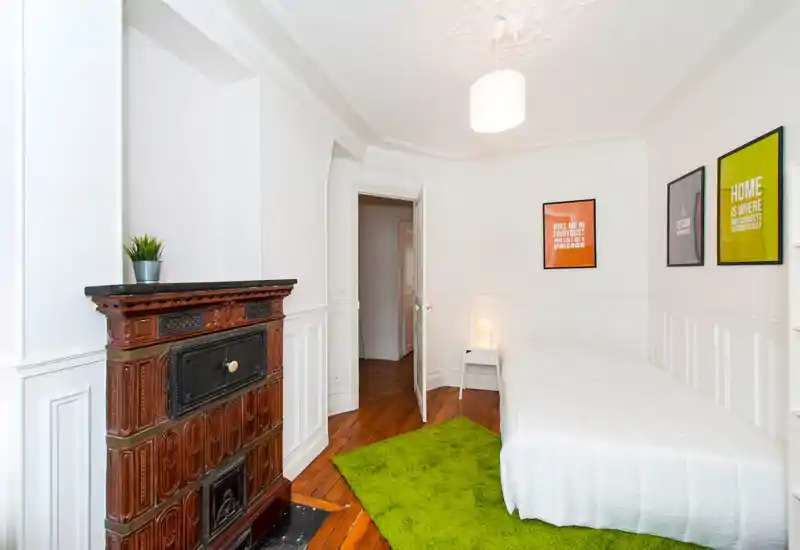 Bright, neatly styled bedroom with a white bed, green rug and decorative vintage stove. Clean lines, wooden floors and framed prints create a cozy, modern feel.