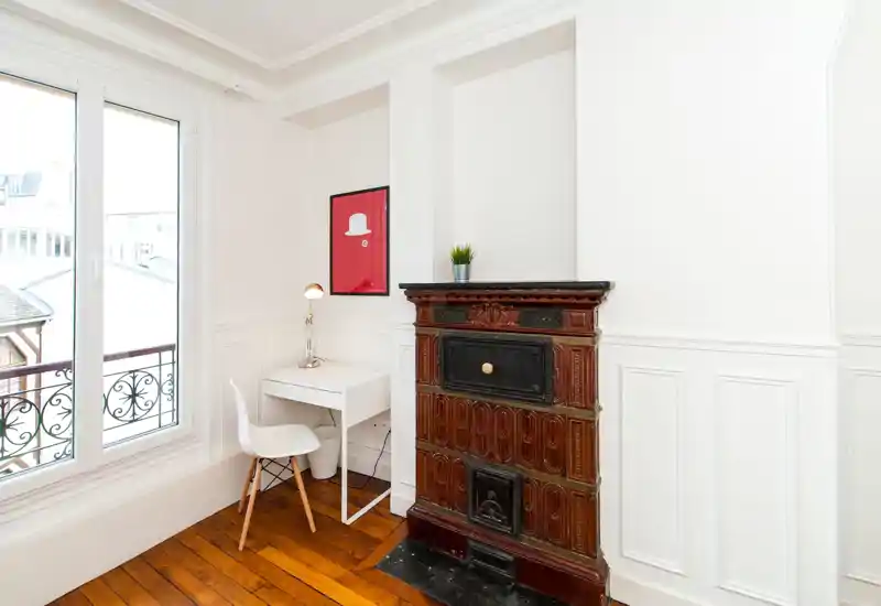Bright, minimalist bedroom corner with a small desk, modern chair and a decorative vintage tiled stove; large window provides abundant natural light.