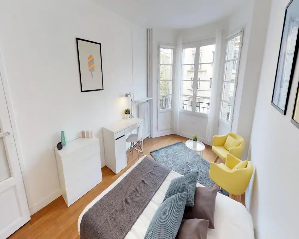Chambre lumineuse et bien mise en scène avec un lit soigné, parquet et un coin salon cosy près de grandes fenêtres laissant entrer beaucoup de lumière naturelle.