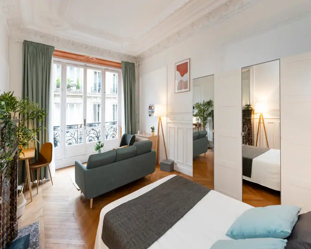 Amplio dormitorio decorado con cama doble en primer plano, zona de estar pequeña y grandes ventanas francesas que proporcionan mucha luz natural.