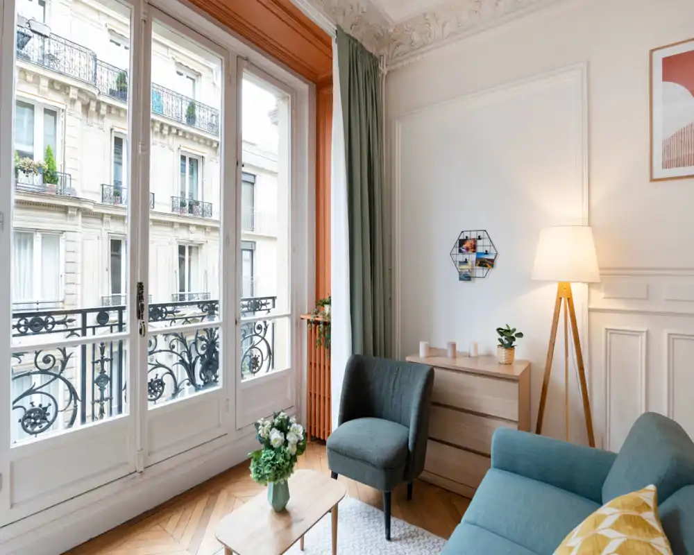 Salón luminoso y elegante con grandes ventanas francesas, butaca y sofá cómodos, lámpara decorativa y mesa de centro de madera — un espacio acogedor y aireado.