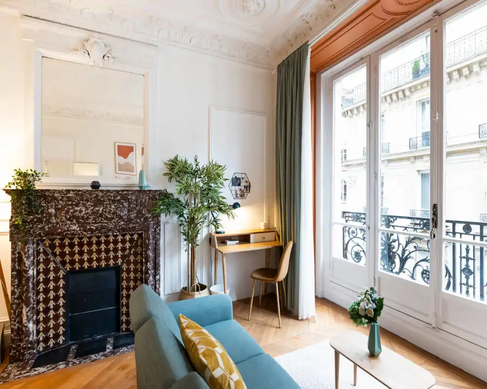 Salón luminoso y elegante con chimenea de mármol decorativa, sofá cómodo con cojines, rincón de escritorio y grandes ventanas de estilo parisino que inundan el espacio de luz.