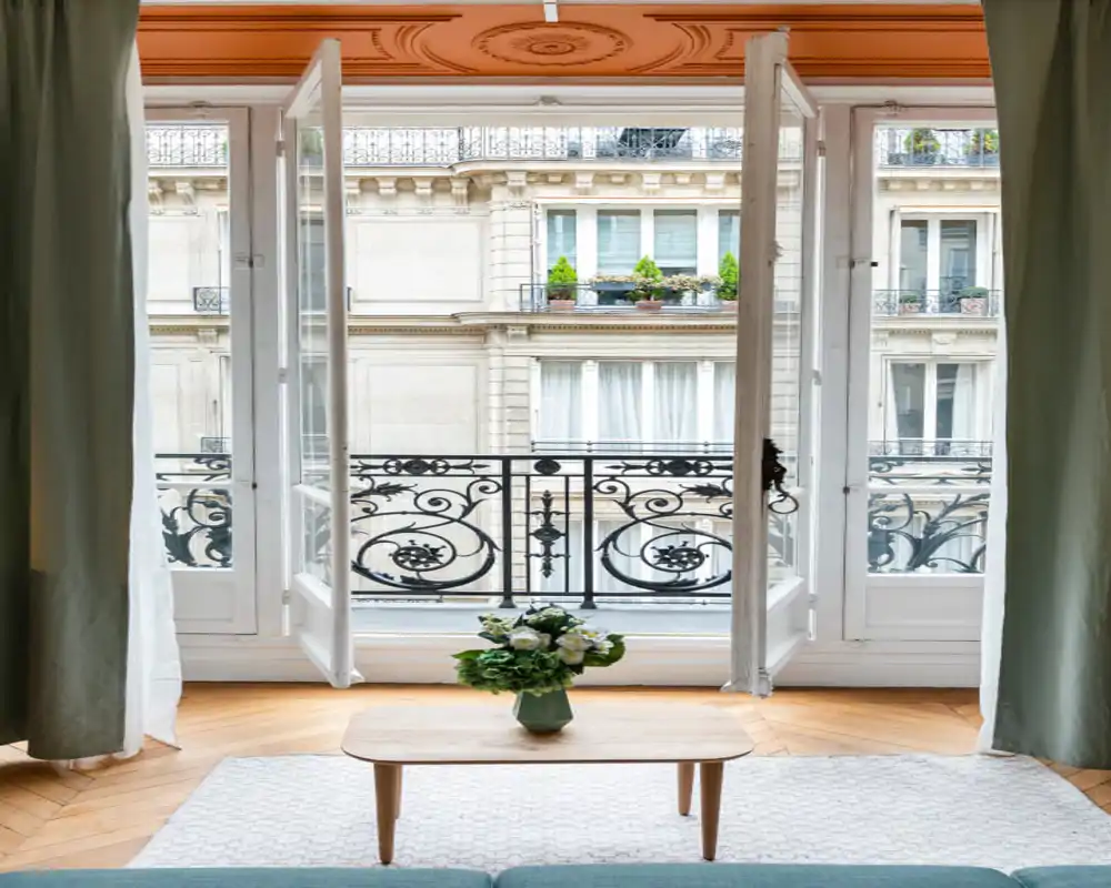Espacio de estar luminoso y elegante con ventanas francesas abiertas, balcón de hierro decorativo, mesita con flores frescas y textiles suaves que crean un ambiente acogedor.