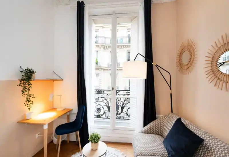 Bright, stylish living corner with a patterned sofa, floor lamp and a small desk by a large window that opens to a balcony — warm textiles and plants add a cozy touch.