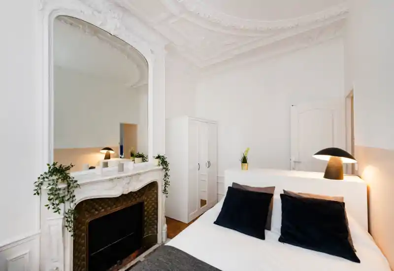 Bright, elegant bedroom with decorative ceiling moldings and a large ornate mirror above a fireplace; comfortable double bed with dark velvet pillows and warm bedside lighting.