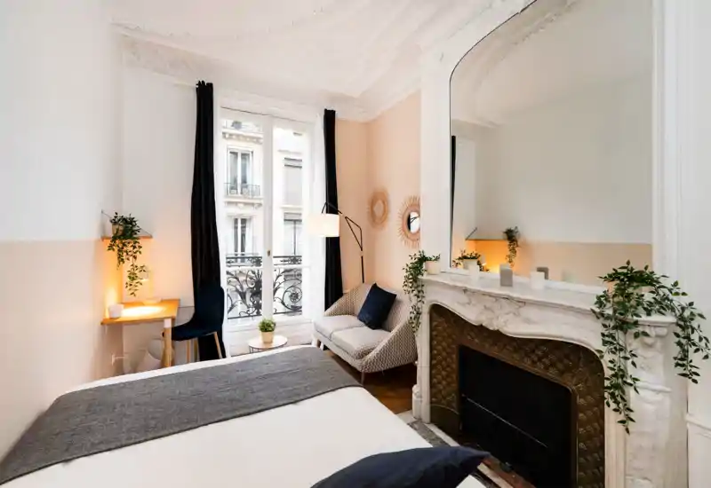 Bright, stylish bedroom with a large bed in the foreground, decorative fireplace with mirror, cozy armchair and desk by a tall window that brings natural light.