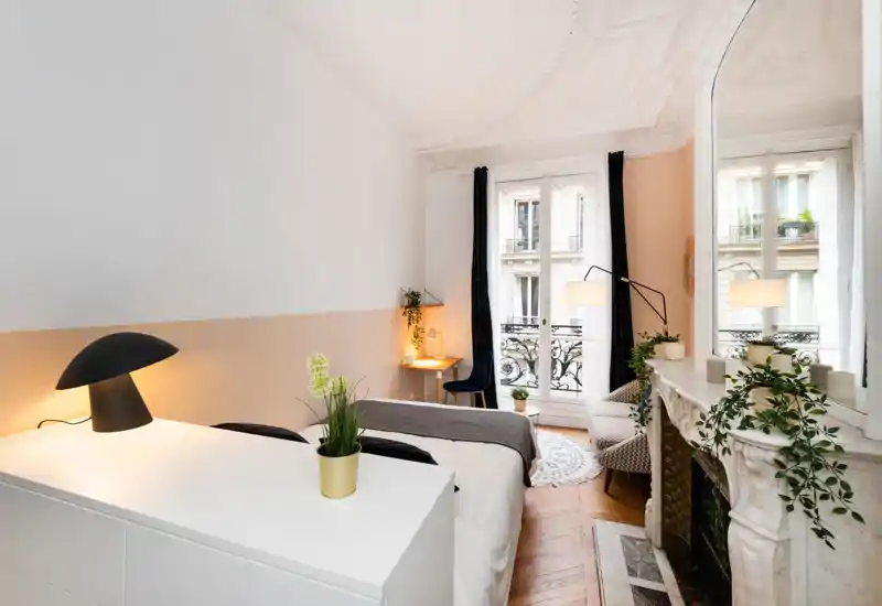 Bright, stylish bedroom with a queen bed, decorative molding and large windows opening to a Parisian balcony. Warm lamps, plants and a cozy armchair create an inviting, well-styled space.