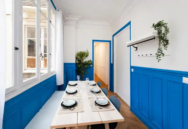 Bright, stylish dining room with a long wooden table set for six, large windows and vivid blue wainscoting that creates a modern, welcoming atmosphere.