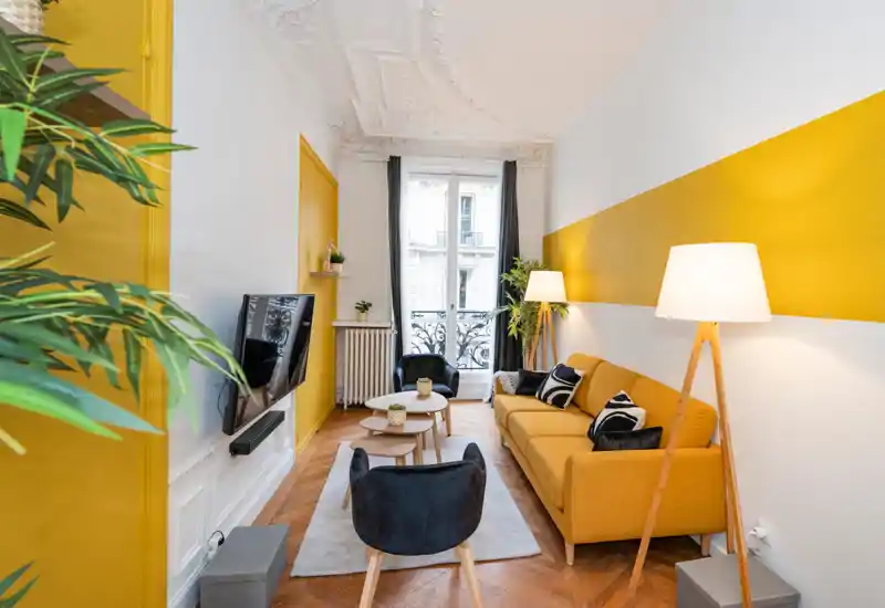 Bright, stylish living room with a mustard-yellow sofa, accent chairs, nested coffee tables and decorative plants; large window provides natural light and elegant architectural details add character.